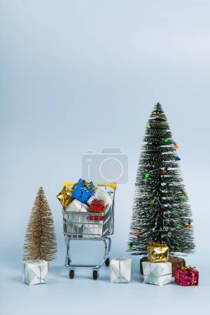 Foto de Conjunto con un caddie lleno de regalos sobre un fondo azul cielo luminoso y árbol de Navidad verde con luces de cuerda. Minimalista, fotografía bodegón de moda. - Imagen libre de derechos