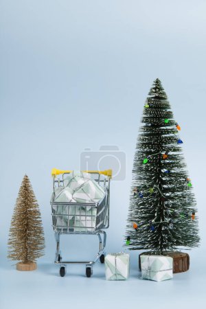 Téléchargez les photos : Ensemble avec un caddie rempli de cadeaux sur un fond bleu ciel lumineux et arbre de Noël vert avec des lumières à ficelle. Photographie de nature morte minimaliste et tendance. - en image libre de droit