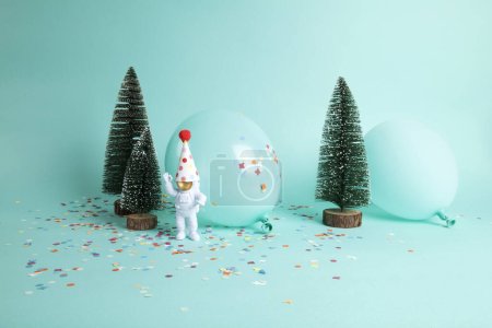 Foto de Un cosmonauta explorando un bosque nevado como un nuevo planeta. Están rodeados de confeti, globos y árbol de Navidad. La composición es de color verde turquesa. Minimalista, fotografía bodegón de moda. - Imagen libre de derechos