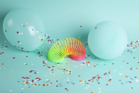 Photo for A plastic rainbow toy and a slinky in front of balloons of the same color as the background and confetti on the ground. The composition is in turquoise-green color palette. - Royalty Free Image