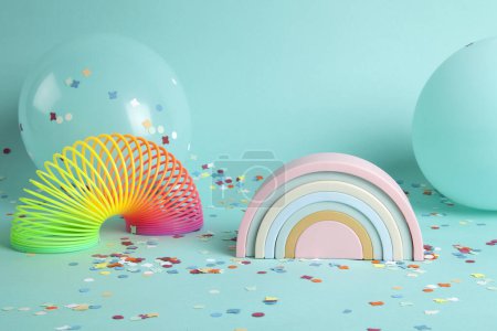 Foto de Un juguete de plástico arco iris y un slinky delante de globos del mismo color que el fondo y confeti en el suelo. La composición es en color verde turquesa paleta. - Imagen libre de derechos