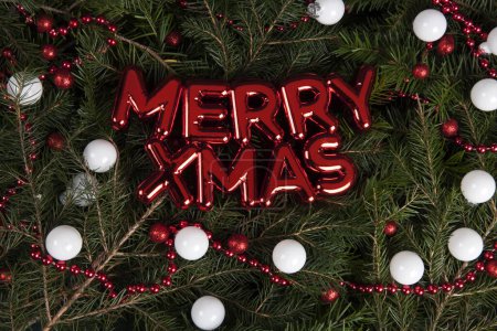 Foto de Feliz Navidad escrito en una decoración de pared de plástico rojo brillante colocado en una pila de ramas de abeto con un marco de guirnalda de perlas de color rojo brillante y muchos adornos de Navidad rojo y blanco. Textura para recurso gráfico - Imagen libre de derechos