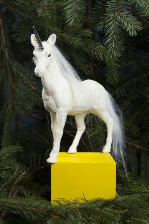 Foto de Un unicornio blanco iridiscente entronizado como una reina del bosque sobre un cubo de madera amarillo. Está rodeada de ramas de abeto, lo que sugiere que está en un bosque mágico.. - Imagen libre de derechos