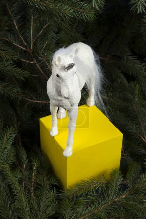 Téléchargez les photos : Une licorne blanche irisée intronisée comme une reine des forêts sur un cube de bois jaune. Elle est entourée de branches de sapin, ce qui suggère qu'elle est dans une forêt magique. - en image libre de droit