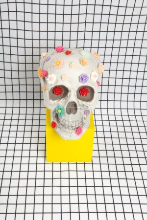 Photo for A skull covered with multiple plastic flowers in different colors placed on a yellow wooden cube on a fabric background with a white squared tile pattern. Metaphor of branches as tomb. vanity as an allegorical representation of the fragility of huma - Royalty Free Image