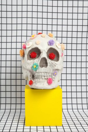 Photo for A skull covered with multiple plastic flowers in different colors placed on a yellow wooden cube on a fabric background with a white squared tile pattern. Metaphor of branches as tomb. vanity as an allegorical representation of the fragility of huma - Royalty Free Image