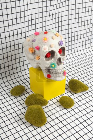 Photo for A skull covered with multiple plastic flowers in different colors placed on a yellow wooden cube and moss rock on a fabric background with a white squared tile pattern. Vanity as an allegorical representation of the fragility of human life - Royalty Free Image