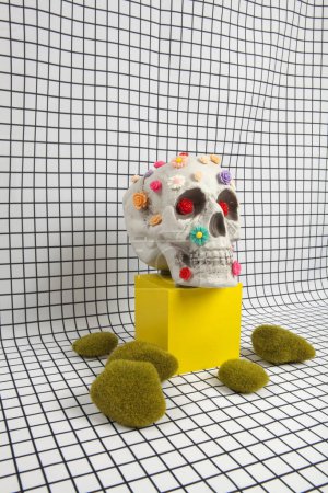 Foto de Un cráneo cubierto de múltiples flores de plástico en diferentes colores colocados en un cubo de madera amarillo y roca de musgo sobre un fondo de tela con un patrón de azulejo cuadrado blanco. Vanidad como representación alegórica de la fragilidad de la vida humana - Imagen libre de derechos