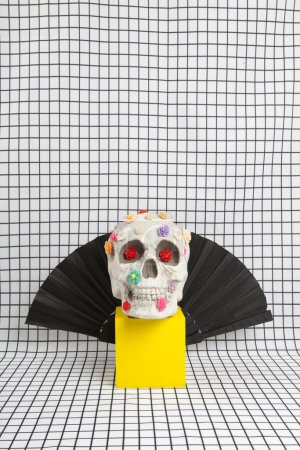 Photo for A skull covered with multiple plastic flowers wearing Count Dracula's high-collared cape placed on a yellow wooden cube on a fabric background with a white squared tile pattern. Vanity as an allegorical representation of the fragility of human life - Royalty Free Image