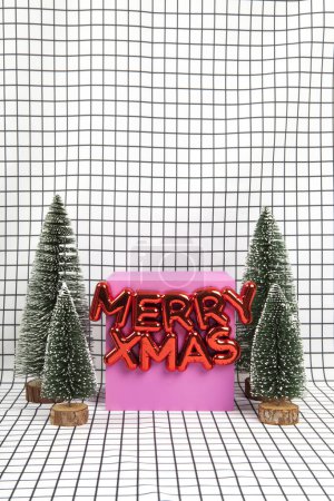 Téléchargez les photos : Un ornement de Noël en plastique rouge brillant avec un texte disant joyeux Noël et une scène de forêt miniature composée de plusieurs petits arbres de Noël et d'un cube jaune sur un fond de grille graphique noir et blanc. photographie de nature morte minimale - en image libre de droit