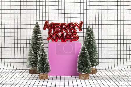 Téléchargez les photos : Un ornement de Noël en plastique rouge brillant avec un texte disant joyeux Noël et une scène de forêt miniature composée de plusieurs petits arbres de Noël et d'un cube jaune sur un fond de grille graphique noir et blanc. photographie de nature morte minimale - en image libre de droit