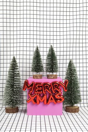 Foto de Un adorno de Navidad de plástico rojo brillante con un texto que dice Feliz Navidad y una escena de bosque en miniatura compuesta por varios árboles de Navidad pequeños y un cubo amarillo sobre un fondo gráfico de rejilla en blanco y negro. fotografía de naturaleza muerta mínima - Imagen libre de derechos