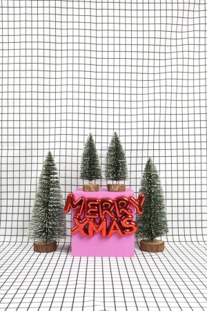 Photo for A glossy red plastic Christmas ornament featuring a text saying merry xmas and a miniature forest scene composed of several small christmas trees and a yellow cube on a graphic black and white grid background. minimal still life photography - Royalty Free Image