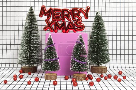 Téléchargez les photos : Un ornement de Noël en plastique rouge brillant avec un texte disant joyeux Noël avec boule de Noël rouge et une scène de forêt miniature composée de plusieurs petits arbres de Noël et un cube jaune sur un fond de grille graphique noir et blanc. minimes encore - en image libre de droit