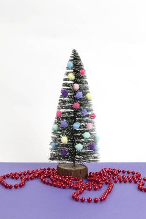 Foto de Árbol de navidad en miniatura decorado con mini guirnaldas y pompones como adornos y una guirnalda de perlas rojas. Fondo violeta y blanco. Bodegón mínimo - Imagen libre de derechos