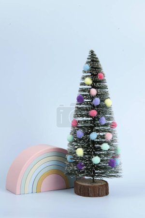 Foto de Árbol de Navidad en miniatura decorado con pompones como bolas con un juguete de plástico arco iris junto a él. Fondo violeta. Fotografía mínima de naturaleza muerta - Imagen libre de derechos