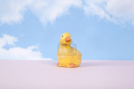 Photo for A plaster figure of a yellow chick on a pink ground and in front of a blue sky background with clouds. Minimal still life photography - Royalty Free Image