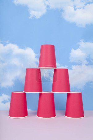 rote Pappbecher, montiert in einer Pyramide, die Erfolg, Gleichgewicht und Stabilität symbolisiert, vor einem blauen Sommerhimmel mit weißen Wolken. Minimale Stillleben-Fotografie.