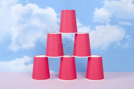 vasos de papel rojo montados en una pirámide que simboliza el éxito, el equilibrio y la estabilidad, frente a un cielo azul de verano con nubes blancas. Fotografía mínima de naturaleza muerta.