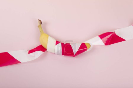 Téléchargez les photos : Une banane mûre, enveloppée dans un ruban de plastique rouge et blanc, retenue prisonnière. Couleur vive et photographie pop art minimale - en image libre de droit