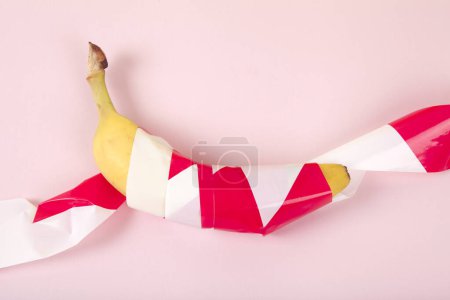 Foto de Un plátano maduro, envuelto en una cinta de plástico roja y blanca, mantenido cautivo. Color vivo y fotografía de arte pop mínima - Imagen libre de derechos