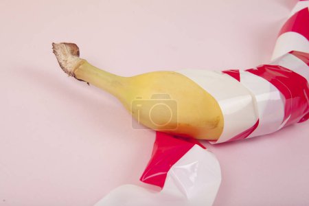 Foto de Un plátano maduro, envuelto en una cinta de plástico roja y blanca, mantenido cautivo. Color vivo y fotografía de arte pop mínima - Imagen libre de derechos