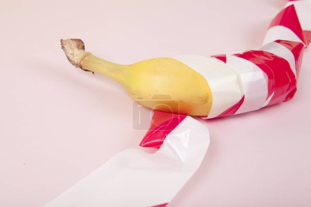 Téléchargez les photos : Une banane mûre, enveloppée dans un ruban de plastique rouge et blanc, retenue prisonnière. Couleur vive et photographie pop art minimale - en image libre de droit