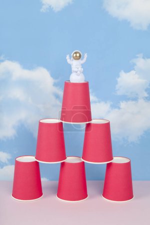 Téléchargez les photos : Une figurine d'astronaute blanc avec casque doré sur une pyramide de gobelets en papier rouge, sur fond de ciel bleu d'été avec des nuages blancs.Couleur vive et photographie pop art minimale - en image libre de droit
