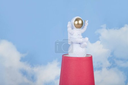 Photo for A white astronaut figurine with gold helmet on a pyramid of red paper cups, set against a background of blue summer sky with white clouds.Vivid color and minimal pop art photography - Royalty Free Image