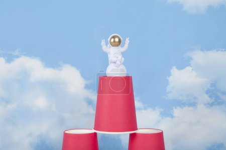 Photo for A white astronaut figurine with gold helmet on a pyramid of red paper cups, set against a background of blue summer sky with white clouds.Vivid color and minimal pop art photography - Royalty Free Image