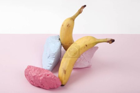 Téléchargez les photos : Une composition scénographique d'une nature morte composée de bananes mûres et de cailloux brillamment peints sur un fond rose et blanc. Couleurs vives et photographie pop art minimale - en image libre de droit