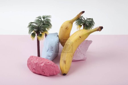 composition scénographique et métaphore d'une île aux palmiers, composée de bananes mûres et de cailloux peints avec éclat sur fond rose et blanc. Couleurs vives et photographie pop art minimale