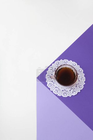 Téléchargez les photos : Un cube violet fait de papier coloré pour créer une illusion d'optique 3D. Sur le dessus se trouve une tasse à café rose vintage avec un napperon en dentelle blanche. Couleurs vives et photographie pop art minimale. - en image libre de droit