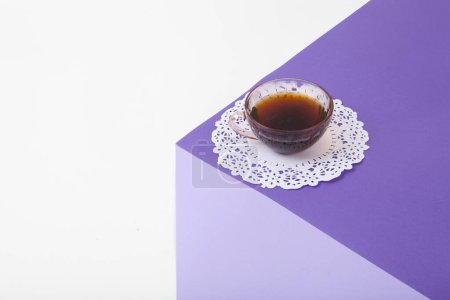 Téléchargez les photos : Un cube violet fait de papier coloré pour créer une illusion d'optique 3D. Sur le dessus se trouve une tasse à café rose vintage avec un napperon en dentelle blanche. Couleurs vives et photographie pop art minimale. - en image libre de droit