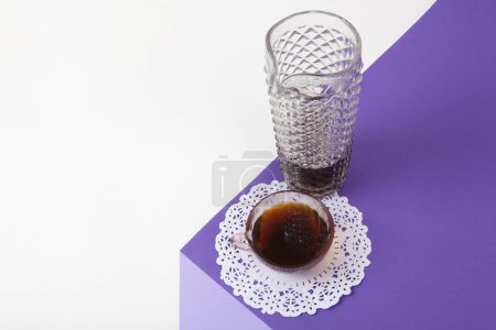 Téléchargez les photos : Un cube violet fait de papier coloré pour créer une illusion d'optique 3D. Sur le dessus se trouve une tasse à café rose vintage avec une dentelle blanche et une cruche en verre d'imitation café, faux cristal. Couleurs vives et photographie pop art minimale. - en image libre de droit