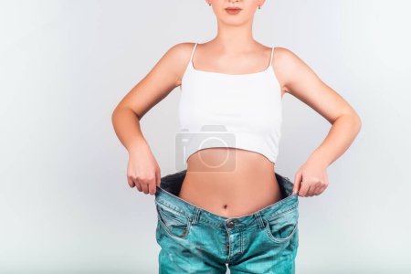 Téléchargez les photos : Diet concept and weight loss. Athletic girl with a beautiful figure in oversize jeans on white background. Body care. - en image libre de droit