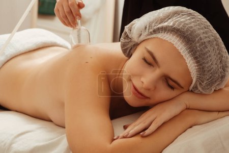 Photo for Young beautiful girl lying on a couch in a cosmetic hat with closed eyes in a spa salon. The masseur is doing wellness procedures, with the help of vacuum jars, body revitalization. High quality photo - Royalty Free Image
