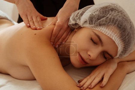 Photo for Young beautiful girl lying on a couch in a cosmetic hat with closed eyes in a spa salon. The masseur is doing wellness procedures, body revitalization. High quality photo - Royalty Free Image