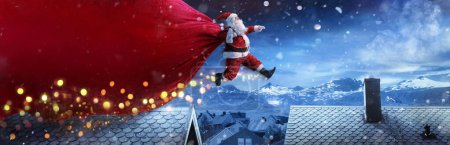 Téléchargez les photos : Père Noël avec le gros sac rouge sautant sur les toits dans un paysage hivernal enneigé - Livraison rapide Présenter - en image libre de droit