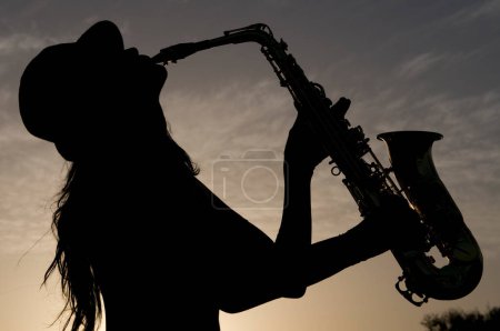 Téléchargez les photos : Silhouette of young woman playing saxophone at sunset in backlight - en image libre de droit