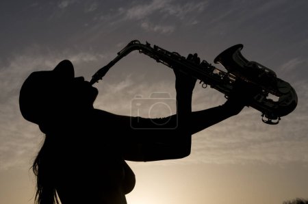 Photo for Silhouette of young woman playing saxophone at sunset in backlight - Royalty Free Image