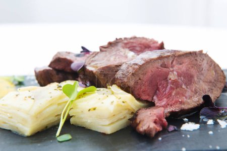 Foto de Carne asada en rodajas con patata y hierbas en un plato negro - Imagen libre de derechos