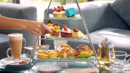 Foto de Woman hand delicately take a slice of cake from a towering display of sweet treats at a fancy restaurant afternoon tea, with cup of hot coffee and tea set nearby. - Imagen libre de derechos