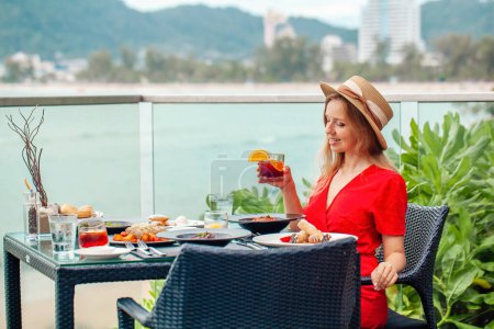 Téléchargez les photos : Une belle fille en vacances se livre à un délicieux dîner avec un cocktail alcoolisé. Restaurant en plein air sur une plage tropicale, bénéficiant de la vue sur la mer. - en image libre de droit