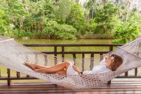 Téléchargez les photos : Femme voyageur embrasse le mode de vie en plein air, couché dans un hamac entouré de montagnes verdoyantes luxuriantes et l'eau du lac vert. Concept de vacances et de détente. - en image libre de droit