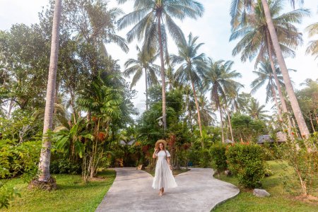 Téléchargez les photos : Femme en robe blanche et chapeau de paille marchant le long d'un sentier tropical en Thaïlande. Concept de loisir et de nature. - en image libre de droit
