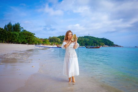Téléchargez les photos : Jeune femme en robe blanche tient coquillage, marche sur la plage. Concept de vacances d'été et de beauté naturelle. - en image libre de droit
