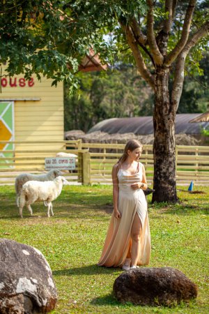 Foto de Mujer sonriente en un vestido, rodeada de naturaleza y ovejas en una granja pacífica. Belleza campestre serena. Mujer en un vestido encantador disfrutando de la idílica vida de granja entre ovejas. - Imagen libre de derechos