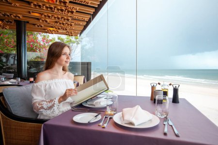 Téléchargez les photos : Élégant jeune adulte au restaurant en bord de mer, tenue décontractée, vue sur l'océan, ambiance gastronomique de luxe, expérience culinaire de voyage. - en image libre de droit