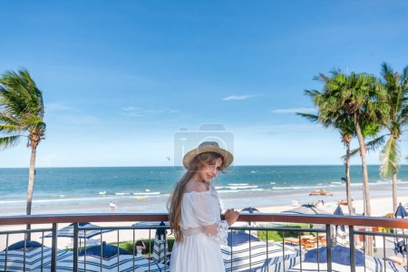 Téléchargez les photos : Femme en robe blanche et chapeau de paille sur balcon donnant sur la plage tropicale avec des palmiers. Style de vie et voyages de luxe. - en image libre de droit
